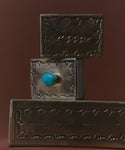 Three rectangular, silver-toned Large Stamped Matchboxes with etched arrow and zigzag designs are stacked. The middle box features a round turquoise stone in front. All are displayed against a solid brown background.