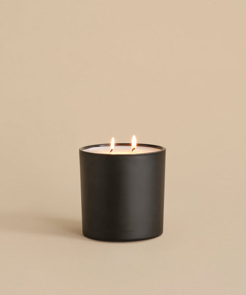 The Large Sun Valley Glass Candle, featuring a hand-poured white two-wick candle in a black matte holder, burns brightly with both flames lit against a plain beige background.