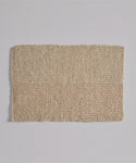The Laurel Bath Mat, a rectangular, woven beige mat made from 100% fique, lies flat on a light gray surface. Its coarse texture with visible fibers and a faint horizontal line gives it an ivory, natural look.