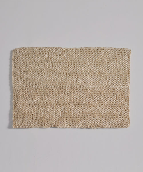 The Laurel Bath Mat, a rectangular, woven beige mat made from 100% fique, lies flat on a light gray surface. Its coarse texture with visible fibers and a faint horizontal line gives it an ivory, natural look.