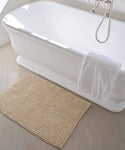 A white freestanding bathtub with a silver faucet and a white towel draped over the side sits next to the Laurel Bath Mat, an ivory mat made from 100% fique, resting on a light wooden floor.