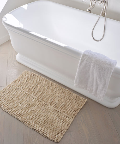 A white freestanding bathtub with a silver faucet and a white towel draped over the side sits next to the Laurel Bath Mat, an ivory mat made from 100% fique, resting on a light wooden floor.