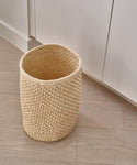 The Laurel Waste Basket in beige, handwoven and natural, rests on a light wood floor beside white cabinets, bringing organic charm to a modern minimalist space.