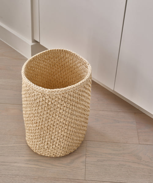 The Laurel Waste Basket in beige, handwoven and natural, rests on a light wood floor beside white cabinets, bringing organic charm to a modern minimalist space.