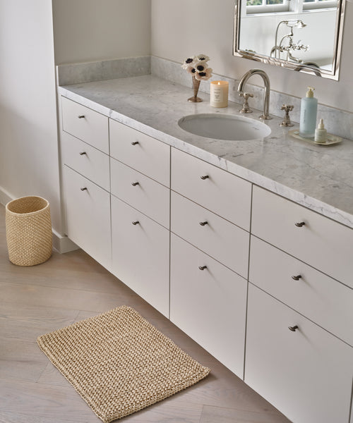 Modern bathroom with a white vanity, marble countertop, round sink, silver faucet, and decor. The Laurel Waste Basket and a matching textured bath mat are on the light wood floor. A mirror hangs above the sink.
