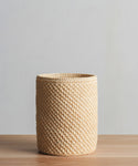 The Laurel Waste Basket, a round handwoven bin crafted from natural fibers, rests on a wooden surface with a plain light gray background.