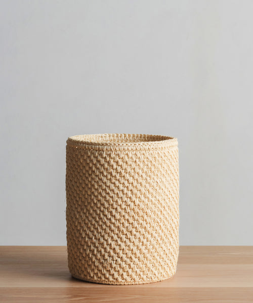 The Laurel Waste Basket, a round handwoven bin crafted from natural fibers, rests on a wooden surface with a plain light gray background.