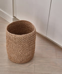 The Laurel Waste Basket, round and handwoven, sits empty on a light wood floor beside white cabinets.