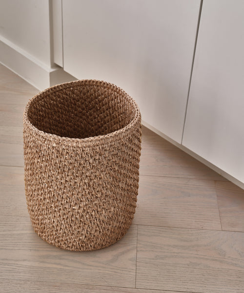 The Laurel Waste Basket, round and handwoven, sits empty on a light wood floor beside white cabinets.