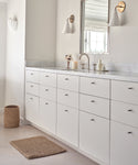A minimalist bathroom with white cabinets, marble countertop, wall-mounted mirror, sconces, a Laurel Waste Basket, and a woven rug on light wood flooring. Toiletries and decor are neatly arranged on the counter.