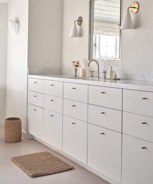A minimalist bathroom with white cabinets, marble countertop, wall-mounted mirror, sconces, a Laurel Waste Basket, and a woven rug on light wood flooring. Toiletries and decor are neatly arranged on the counter.