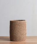 The Laurel Waste Basket, a round handwoven piece in light brown fique, sits on a wooden surface with a plain light gray background.