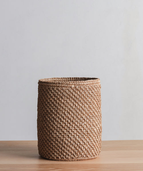 The Laurel Waste Basket, a round handwoven piece in light brown fique, sits on a wooden surface with a plain light gray background.