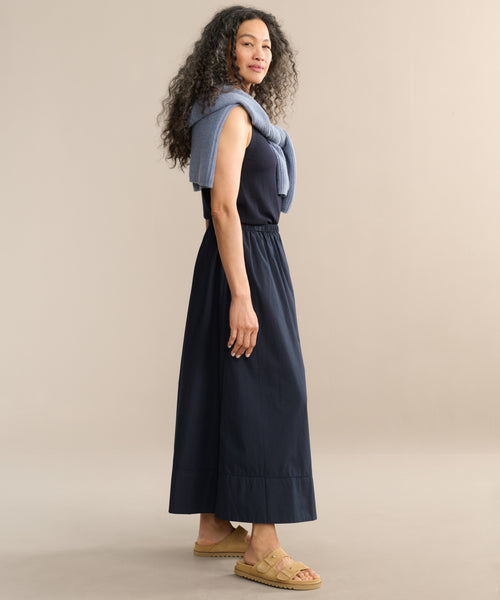 A woman stands sideways against a neutral background, wearing a sleeveless navy top, the Lauren Skirt, tan sandals, and a light blue sweater draped over her shoulders.
