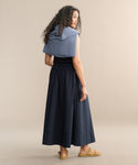 A woman with long curly hair stands facing away against a plain beige background, wearing a sleeveless black top, the Lauren Skirt in navy, tan sandals, and a blue sweater draped over her shoulders.