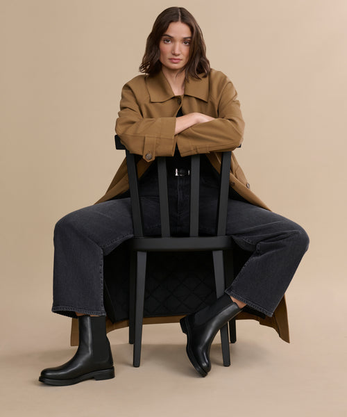 A woman wearing a tan trench coat, black jeans, and Leather Alden Boots sits backward on a black chair against a beige background, her arms resting on the chair’s back and legs apart.