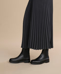 A person wearing a long, dark pleated skirt and Leather Alden Boots stands on a beige background. Only the lower half of their legs and feet are visible.