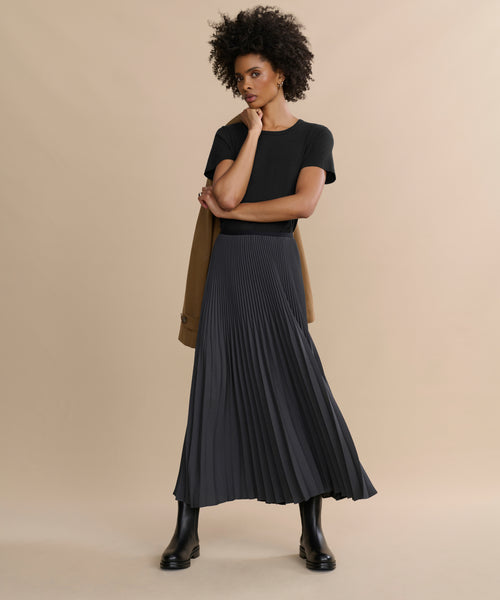 A woman poses against a beige background in a black short-sleeve top, long pleated black skirt, and tan coat over her shoulder. She completes the look with sleek Leather Alden Boots and rests one hand thoughtfully on her chin.