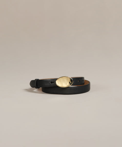 The Leather Audrey Belt, crafted from black premium calf leather with a shiny oval-shaped brass buckle, is coiled on a plain light beige background.