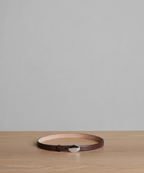 The Leather Audrey Belt, crafted in Italy from brown calf leather, is displayed in a circular shape on a light wooden surface with a gray background.