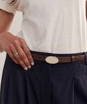 Wearing a cream shirt and navy trousers, the individual showcases the Leather Audrey Belt—an elegant Italian-made calf leather belt with an oval buckle—while resting one hand on their waistband and rings on every finger.
