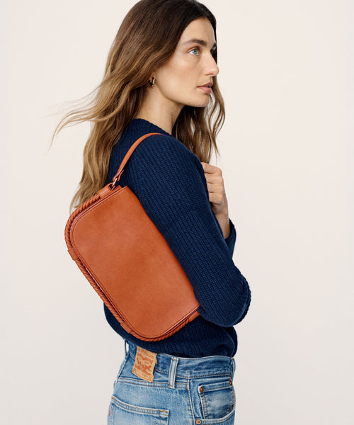 A woman with long wavy brown hair, in a navy sweater and light blue jeans, carries the Brooke Shoulder Bag in Vachetta leather with woven trim, made in Italy, standing against a plain light background.