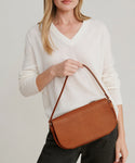A woman in a white v-neck sweater and dark pants holds the Brooke Shoulder Bag, made from Vachetta leather, at her waist against a plain light background. Made in Italy.