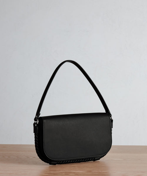The Brooke Shoulder Bag features a single strap and classic rectangular silhouette with rounded edges in black Vachetta leather. Shown on a light wooden surface with a plain, light background.