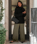 A woman with shoulder-length dark hair and red lipstick stands by a black door, wearing a black turtleneck, olive green wide-leg pants, black heels, and carrying the Brooke Shoulder Bag.