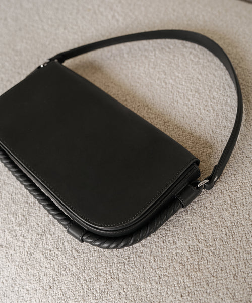 The Brooke Shoulder Bag is a black leather bag with a smooth finish and braided trim, displayed on a light fabric surface. Expertly crafted in Italy, this elegant accessory showcases timeless style and quality.