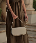 Outdoors, a person in a sleeveless brown dress holds the Brooke Shoulder Bag in beige Vachetta leather. Stone steps and lush greenery complete the background.