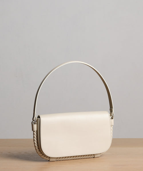 The Brooke Shoulder Bag showcases a minimalist design with a cream finish, curved top handle, and flap closure, plus subtle rope detailing. Crafted from premium Vachetta leather in Italy for timeless elegance.