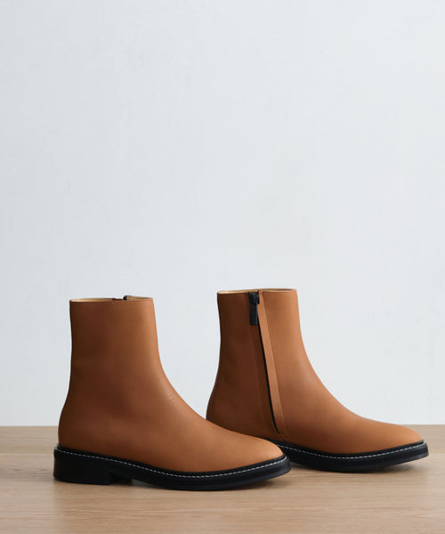 The Blake Ankle Boot is a tan leather ankle boot with black soles and white stitching, displayed on wood. One boot is zipped up, while the other shows the side zipper, set against a plain light background.