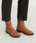 A person stands on a light-colored carpet wearing black pants and the Blake Ankle Boot—brown leather with black soles and white stitching. This stylish zip-up ankle boot adds a sophisticated touch to the look.