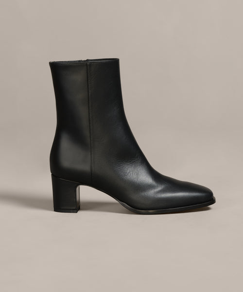 The Leather Bobby Boot, crafted from black Italian leather with a mid-height block heel, rounded square toe, and side zipper, is shown against a neutral beige background.