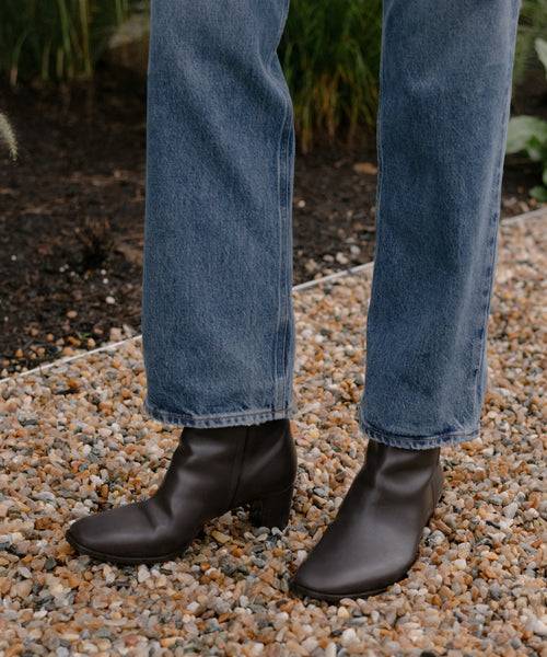 A person in blue jeans wears the versatile Leather Bobby Boot for women, standing on a gravel path with green plants nearby.