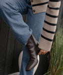 A person in blue jeans and a beige striped sweater adjusts the versatile Leather Bobby Boot on their foot, with a dark wooden wall and tall green grass in the background.