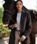 A woman in the Leather Brooke Blazer and a white shirt stands beside a dark brown horse, holding its bridle. Outdoors with greenery behind her, she looks confidently at the camera.
