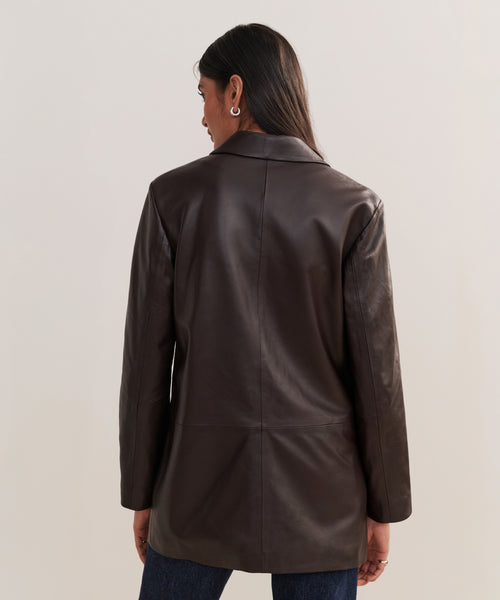 A person with long dark hair and hoop earrings models the Leather Brooke Blazer, a single-breasted leather blazer with an elongated silhouette, paired with dark jeans, posing with their back to the camera against a neutral background.