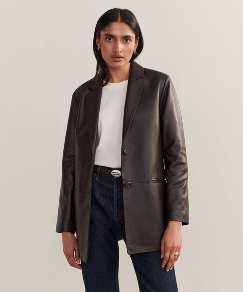 A woman poses against a plain background wearing the Leather Brooke Blazer in dark brown over a light gray top and dark blue jeans, her right hand by her side and left hand in her pocket, highlighting an elongated silhouette.