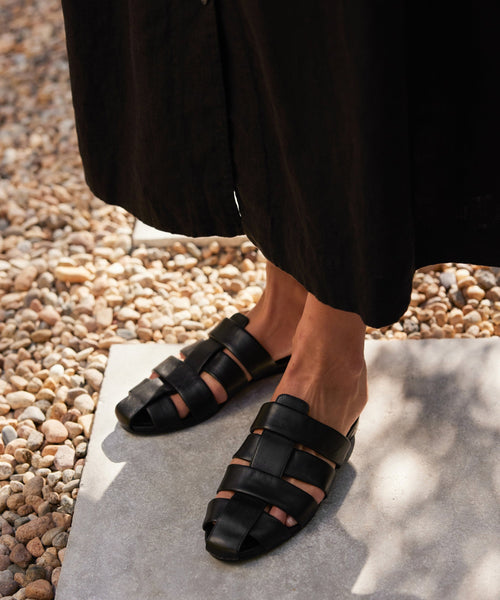 A person stands on a stone path in a long, dark skirt, showing their feet in black Leather Camilla Mules with woven leather, combining comfort and timeless style.