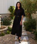 A woman with long dark hair stands on a stone path in a garden, wearing a black belted button-down dress and Leather Camilla Mules. She looks at the camera, surrounded by greenery and a small fountain.