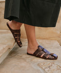 A person wearing a dark skirt stands on stone steps, showing their feet in comfortable Leather Camilla Mules—brown leather sandals with multiple straps inspired by classic fisherman styles.