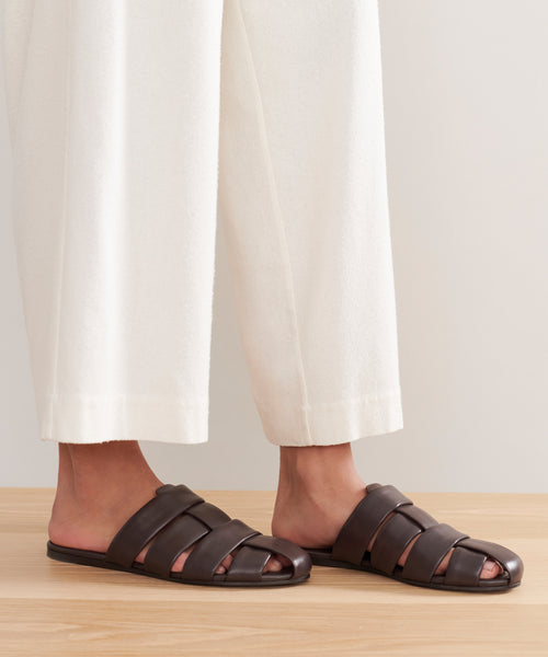 A person wears Leather Camilla Mules—comfortable slides with wide straps—paired with loose white pants, standing on a light wooden floor against a neutral background.