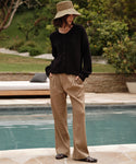 A person stands by a pool wearing a wide-brimmed straw hat, a black button-up sweater, loose beige trousers, and the Leather Camilla Mule, with greenery and a lounge chair in the background.