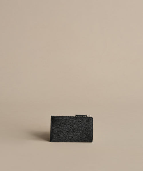 The Leather Card Holder, crafted in small black textured leather, stands upright on a beige surface with a minimalist background—perfect for keeping your travel cards organized on any journey.