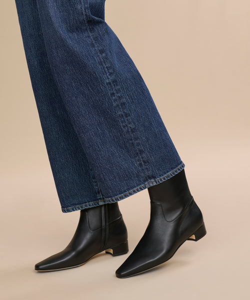 A person stands against a beige background wearing dark blue wide-leg jeans and the Leather Carmine Boot, styled in black leather with a low heel.