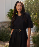 A woman with long dark hair stands outdoors, wearing a short-sleeved black button-up dress and the Classic Belt made in Italy. She faces the camera, with greenery and a stone wall behind her.