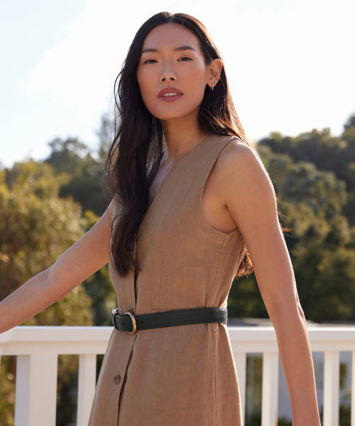 On a sunny day, a woman with long dark hair is outdoors by a white railing, surrounded by greenery. She wears a sleeveless tan dress styled with the Classic Belt.