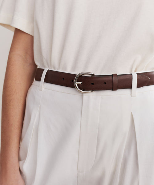 A person wears an off-white short-sleeve top tucked into high-waisted, pleated white pants, finished with the Classic Belt—crafted in Italy from nappa leather and featuring a silver buckle.